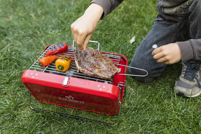 Barbecue à charbon pliable portatif Kikirikit rouge – Mise en situation sur l'herbe du barbecue ouvert , avec une personne en train d’y cuire des aliments.