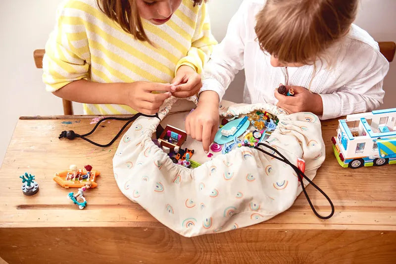 Mini sac de rangement et tapis de jeux motif arc-en-ciel de Playandgo - vue du sac ouvert avec des jeux sur un banc d'école et deux enfants qui jouent 