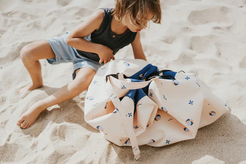 Tapis de jeu et sac de rangement d’extérieur Play and Go – motif ballons – vue du sac en version rangement, posé sur le sable avec un enfant à côté